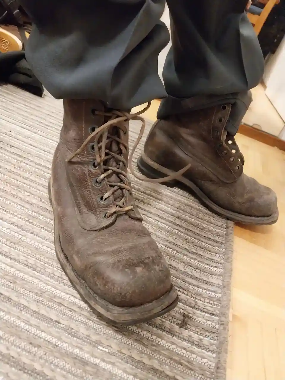 A close-up shot of a person's worn, dirty brown work boots standing on a striped rug over hardwood flooring. The person is…