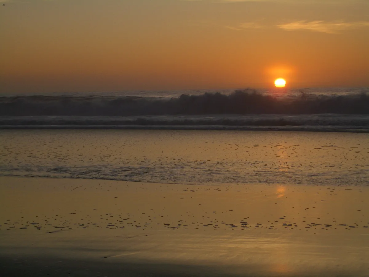 A serene beach sunset with the sun low on the horizon, casting orange and yellow light across the water and wet sand. Waves…