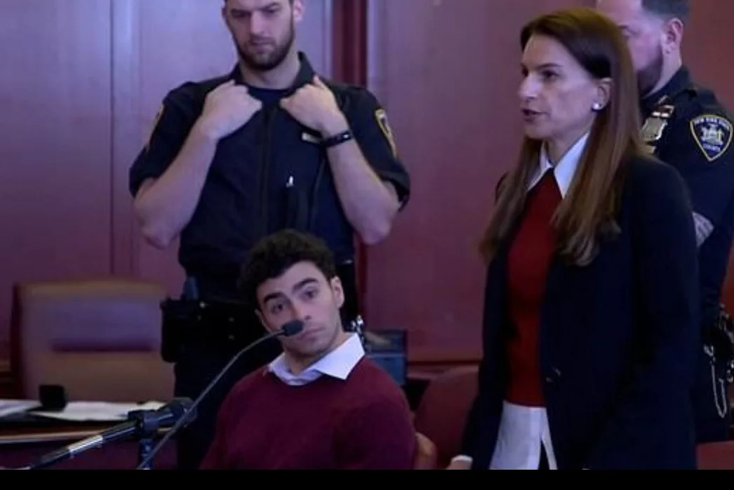 A young man sits in a courtroom with a microphone, looking confused or overwhelmed. Behind him, a police officer adjusts his…