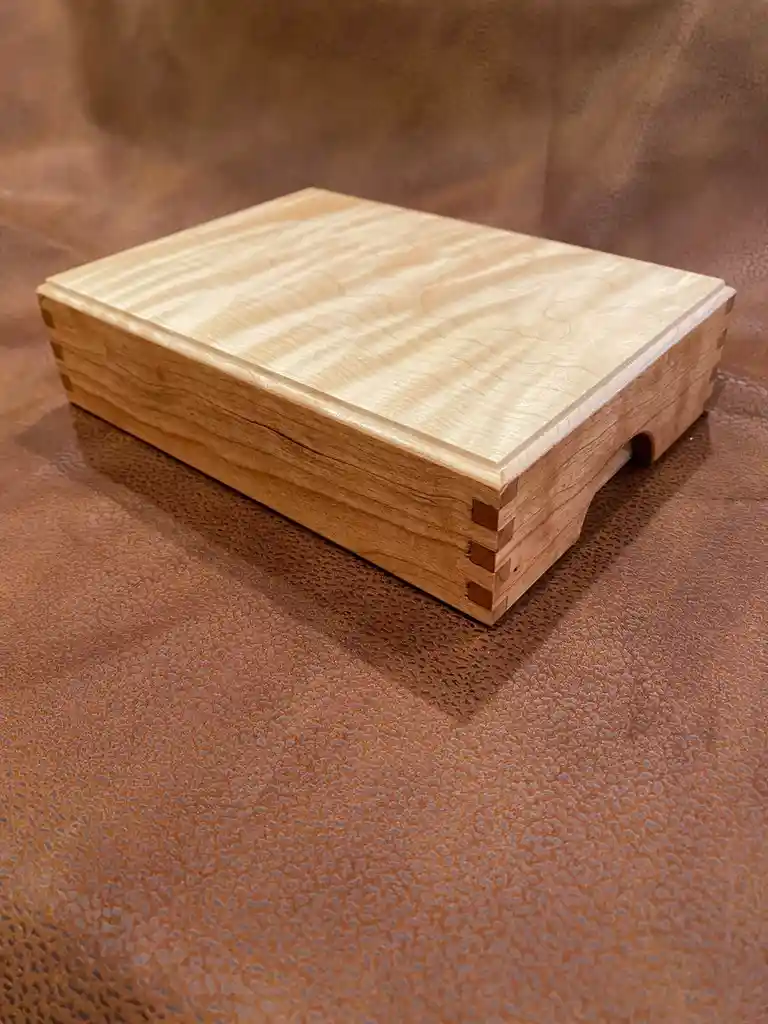 A handcrafted wooden box with visible dovetail joints sits on a brown textured leather surface. The box has a light-colored…