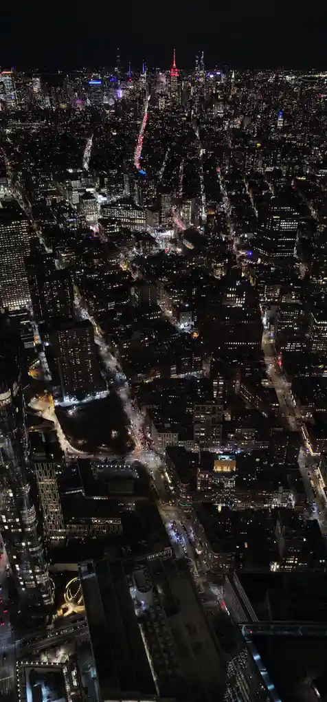 An aerial night photograph of a densely lit cityscape, likely New York City, showing countless illuminated skyscrapers and…