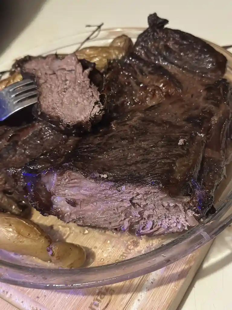 A close-up photo shows a large, darkly seared piece of meat on a glass plate with roasted potatoes, partially sliced to…