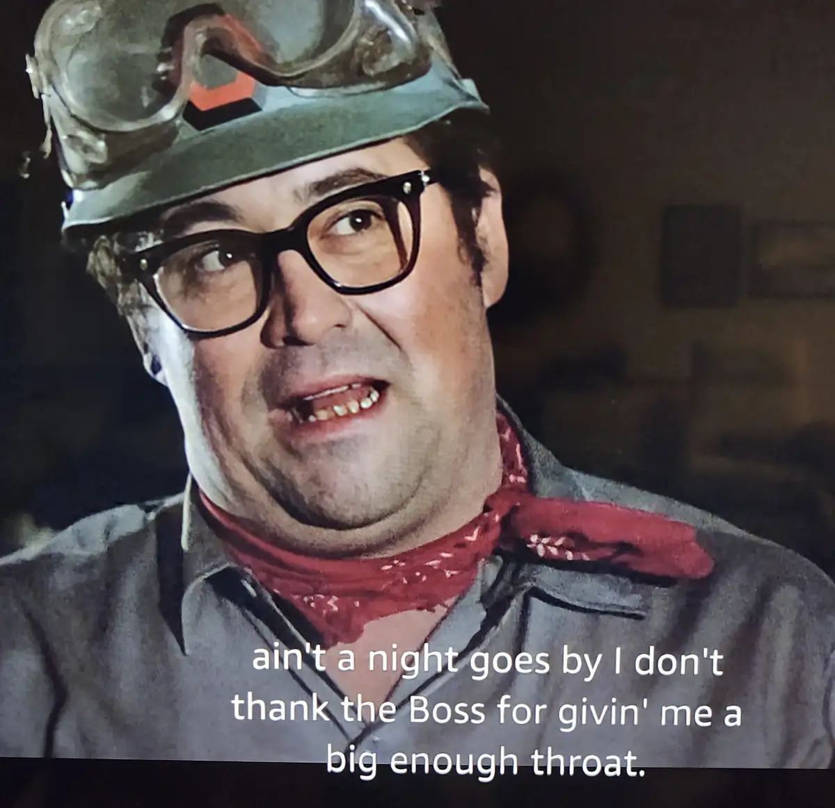 A man wearing a miner's helmet with a lamp, thick black-rimmed glasses, and a red bandana around his neck, smiles while…
