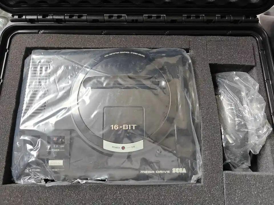 A Sega Mega Drive console, still sealed in plastic, sits in its foam-lined case next to a wrapped controller. The console's…