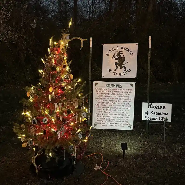 A decorated Christmas tree with lights and antlers stands next to three signs. The top sign reads 'KREWE OF KRAMPUS BE NICE…