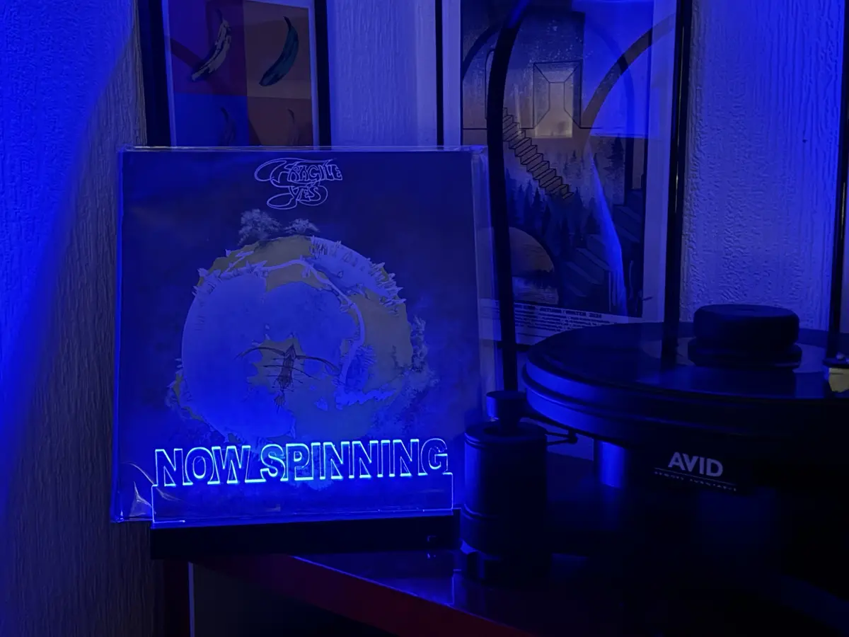 A vinyl record player with an 'AVID' brand turntable sits next to a glowing neon sign displaying 'NOW SPINNING'. The sign is…