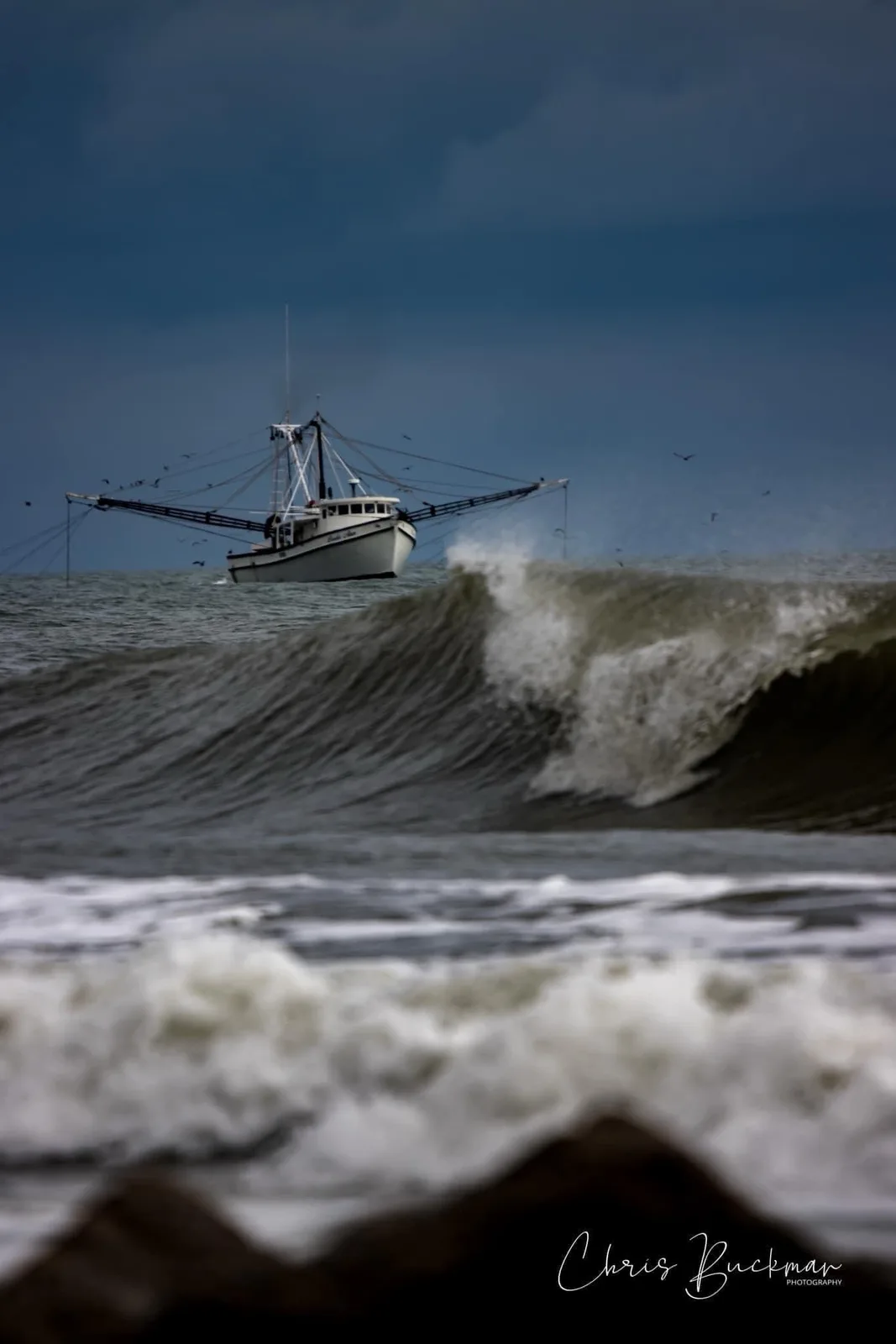 A white shrimp boat navigates choppy, dark ocean waters under a stormy sky, with a large wave cresting in the foreground…