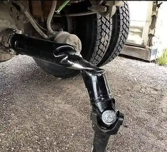 A close-up photo shows a shiny black tow hitch on a vehicle, bent into a distinct hook shape, with a tire and gravel ground…