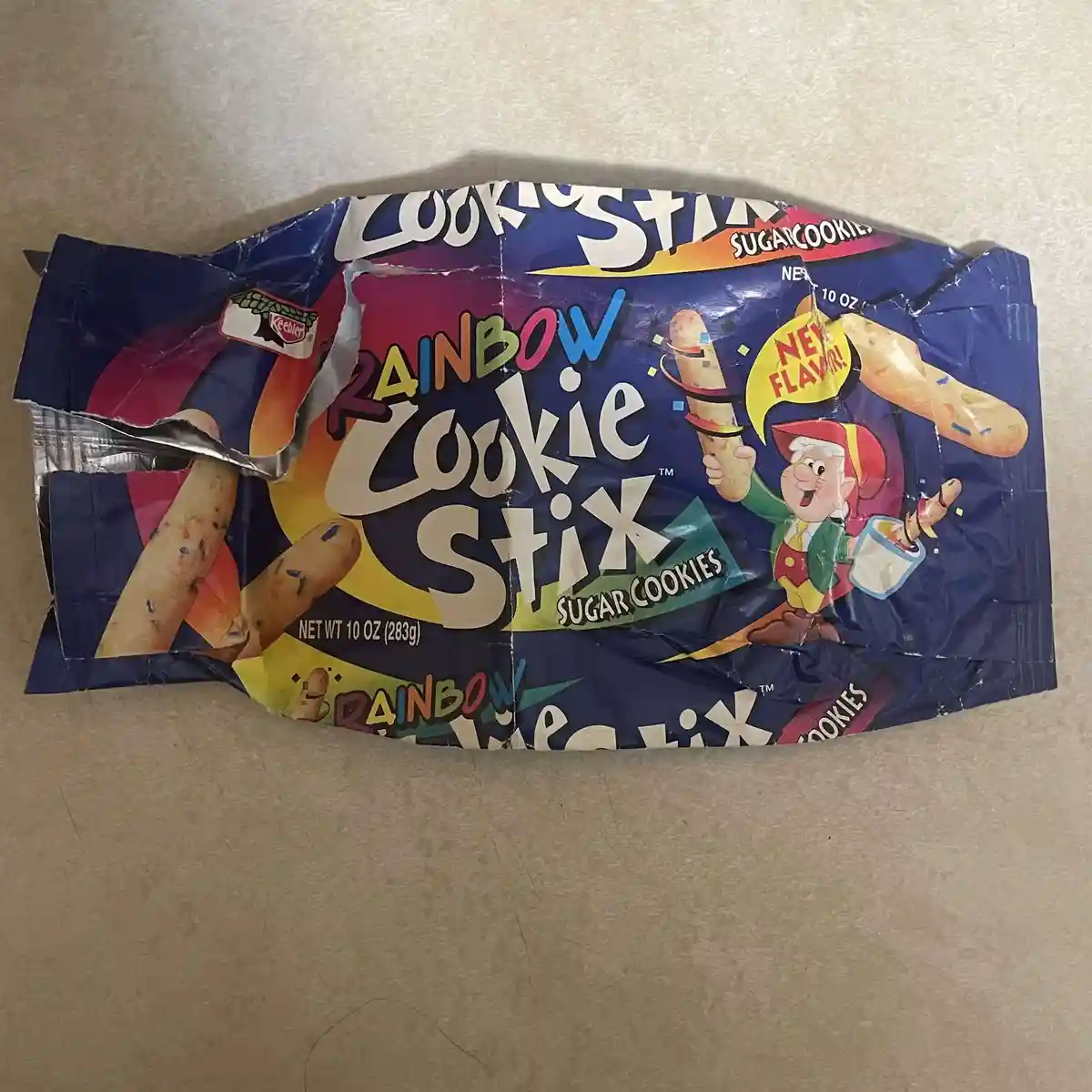A crumpled, torn Keebler Rainbow Cookie Stix package lies on a beige surface, featuring a cartoon elf holding cookie sticks…