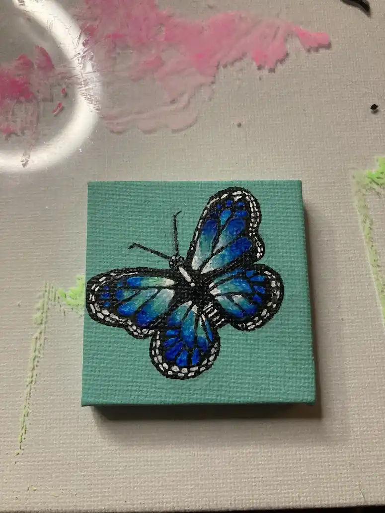 A small square canvas with a painted blue butterfly on a teal background, surrounded by paint splatters and a circular light…