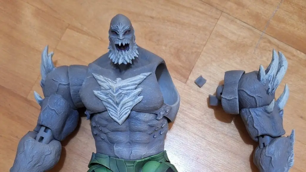 A gray, muscular action figure with a monstrous face and green pants lies on a wooden floor, its right arm detached and…