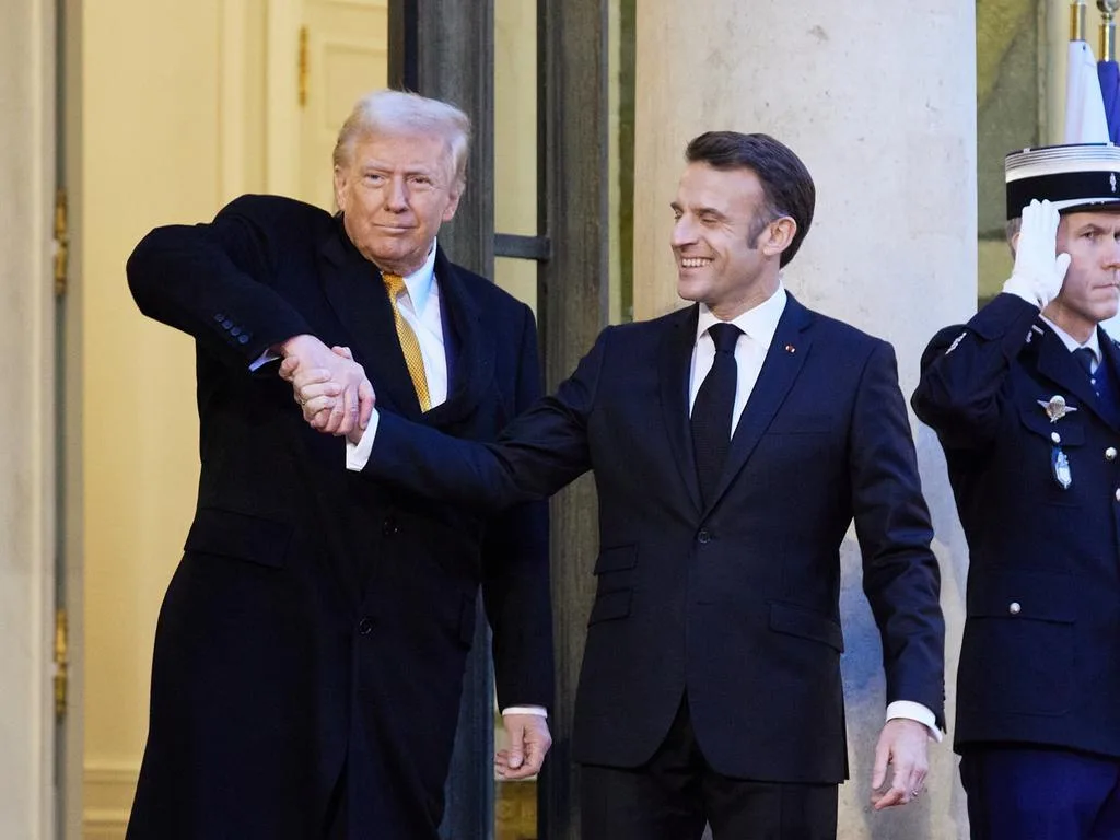 two men in suits shake hands, one smiling broadly while the other has a stern expression and his arm is awkwardly…