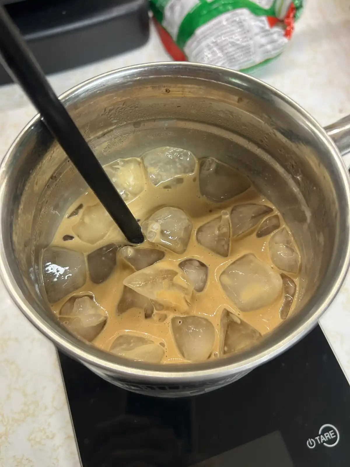 A stainless steel pot sits on a black induction cooktop, filled with a light brown iced coffee beverage and numerous ice…