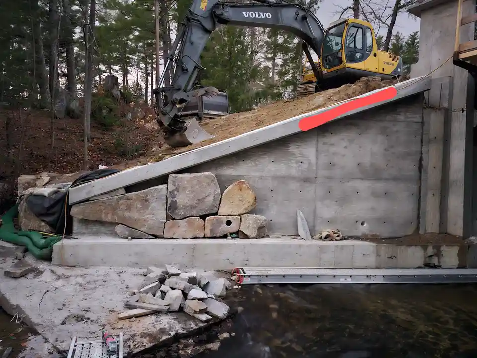 A construction site features a yellow Volvo excavator digging near a concrete retaining wall next to a body of water. A red…