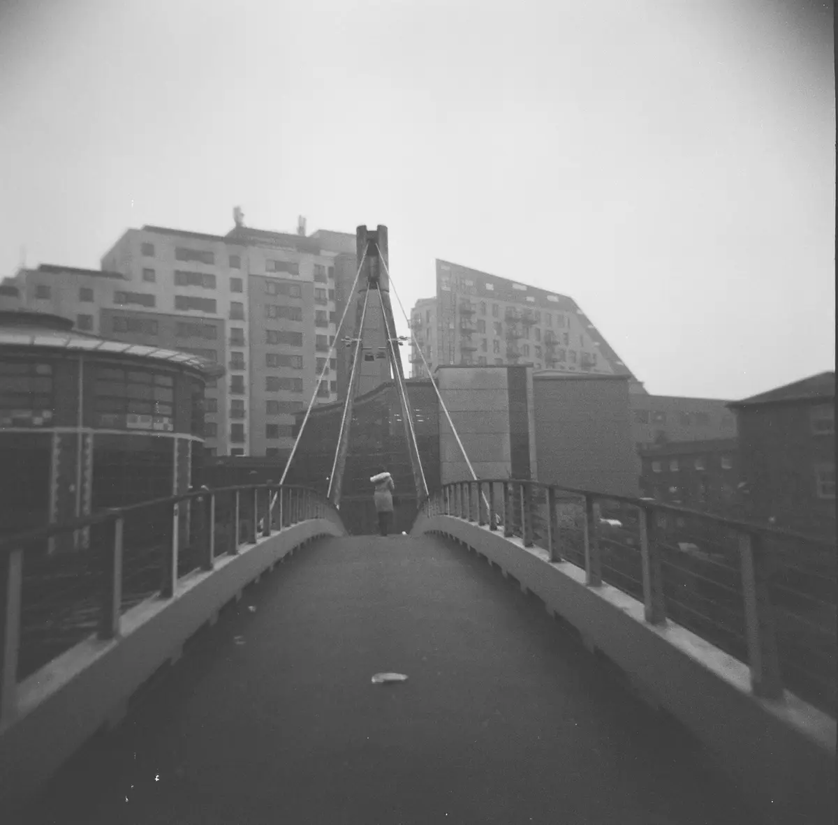 A black-and-white photo shows a lone person walking away on a modern pedestrian bridge, with city buildings in the…