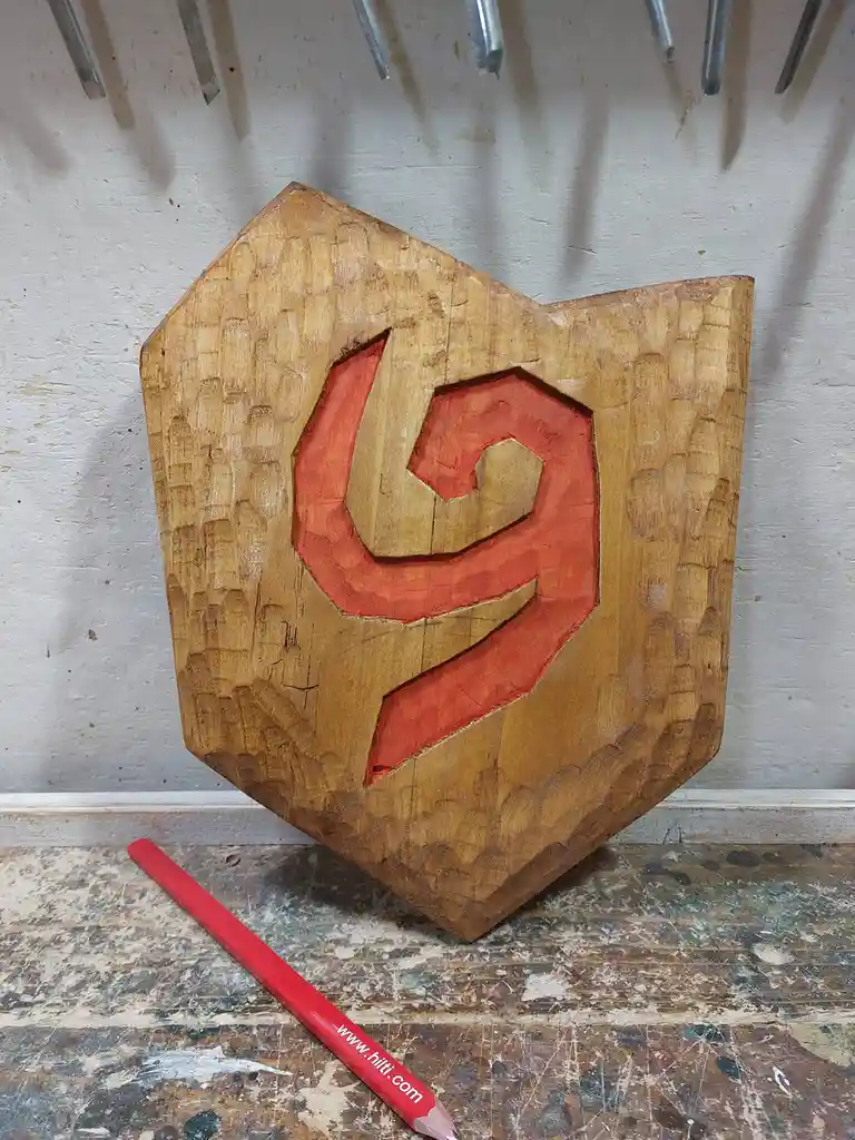 A wooden carving of the red Hilti logo, shaped like a shield, sits on a worn workbench with a red pencil bearing the Hilti…