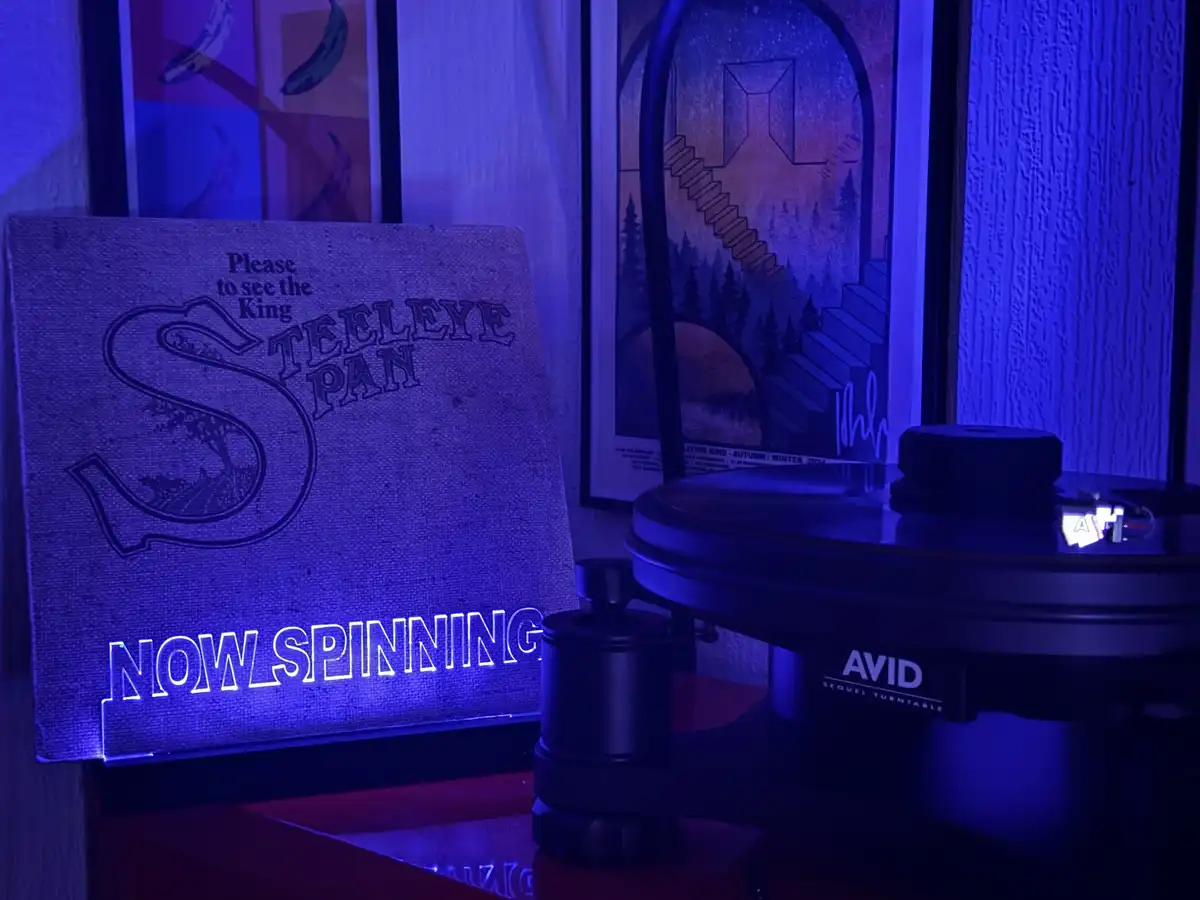 A vinyl record player, branded 'AVID SEQUEL TURNTABLE', sits under blue lighting next to a canvas sign that reads 'Please to…