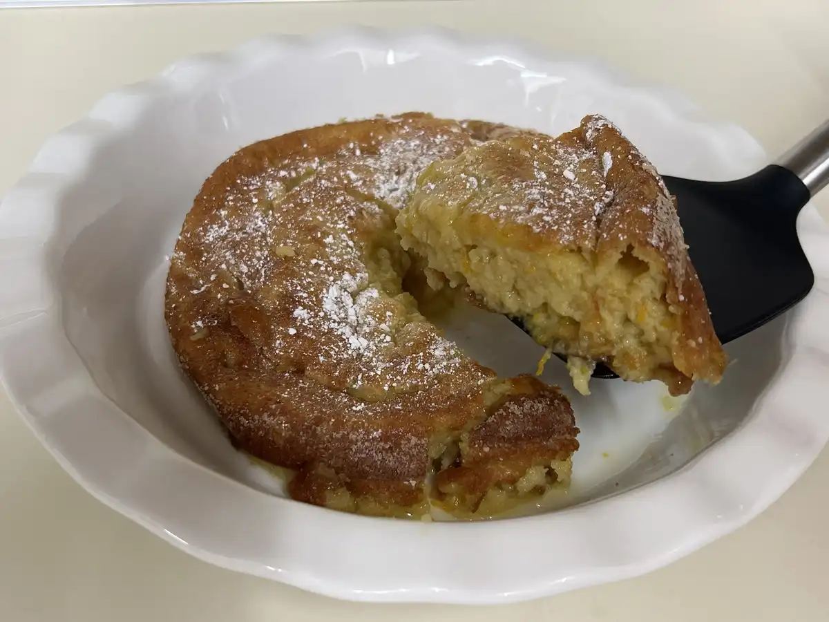 A close-up photo shows a golden-brown baked dessert, likely a clafoutis or custard-based cake, in a white scalloped ceramic…