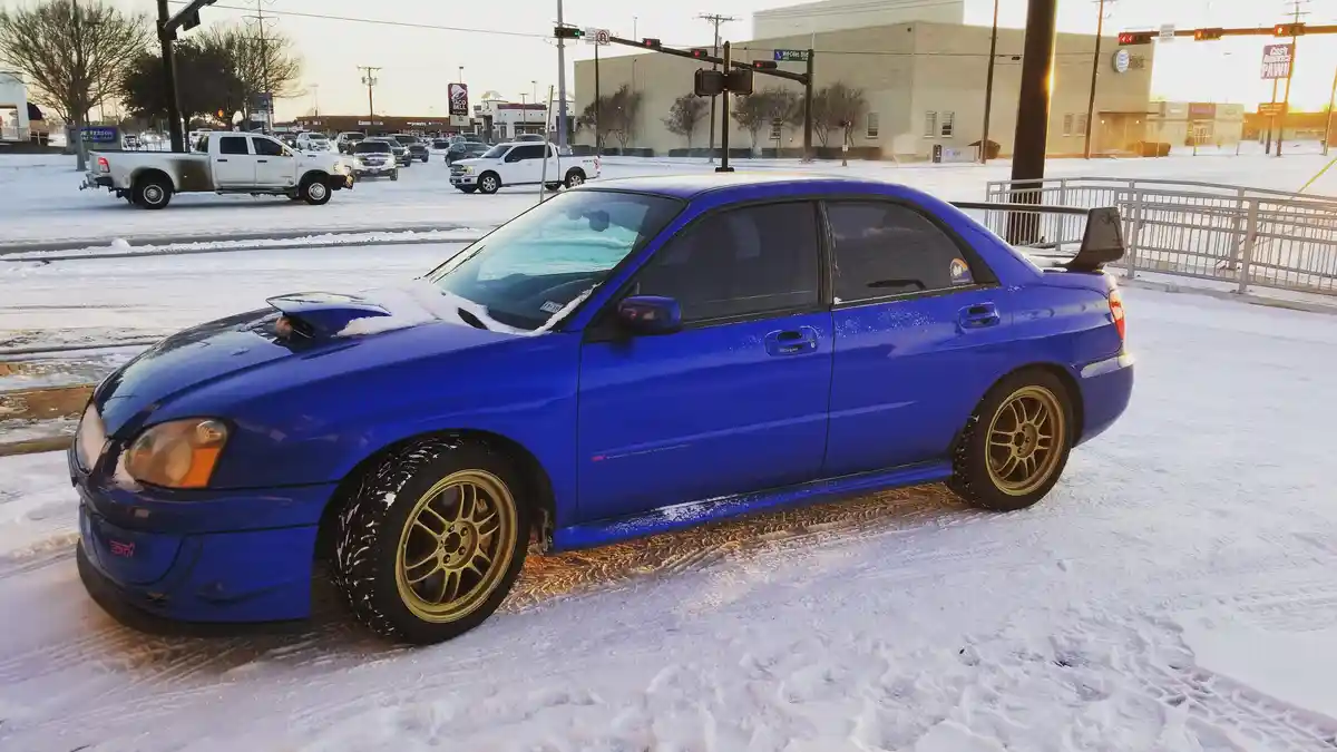A vibrant blue Subaru Impreza WRX STI is parked in a snowy parking lot, its hood and roof dusted with snow, while gold…