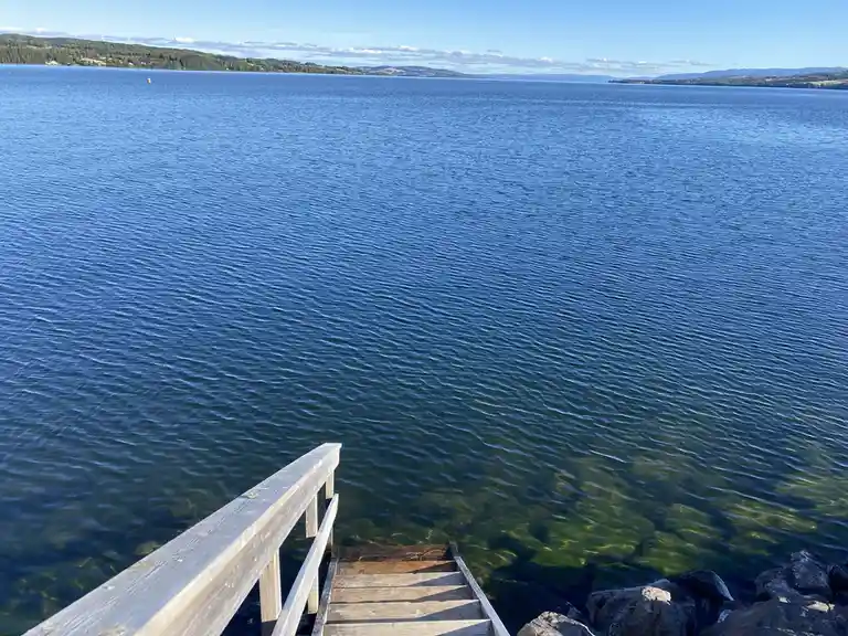 A wooden dock with stairs extends into a calm, blue lake under a clear sky, with distant tree-covered hills on the horizon…