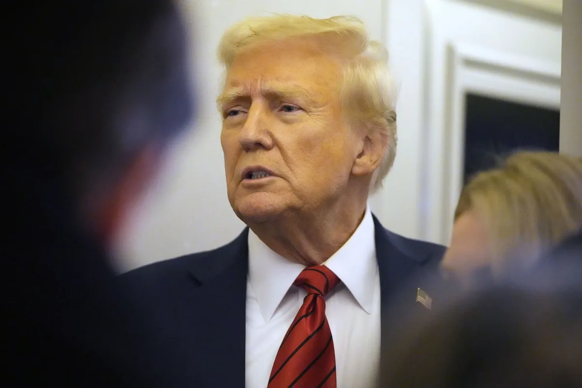 a photo of Donald Trump in a dark suit and red striped tie, looking slightly confused or exasperated while speaking, with…