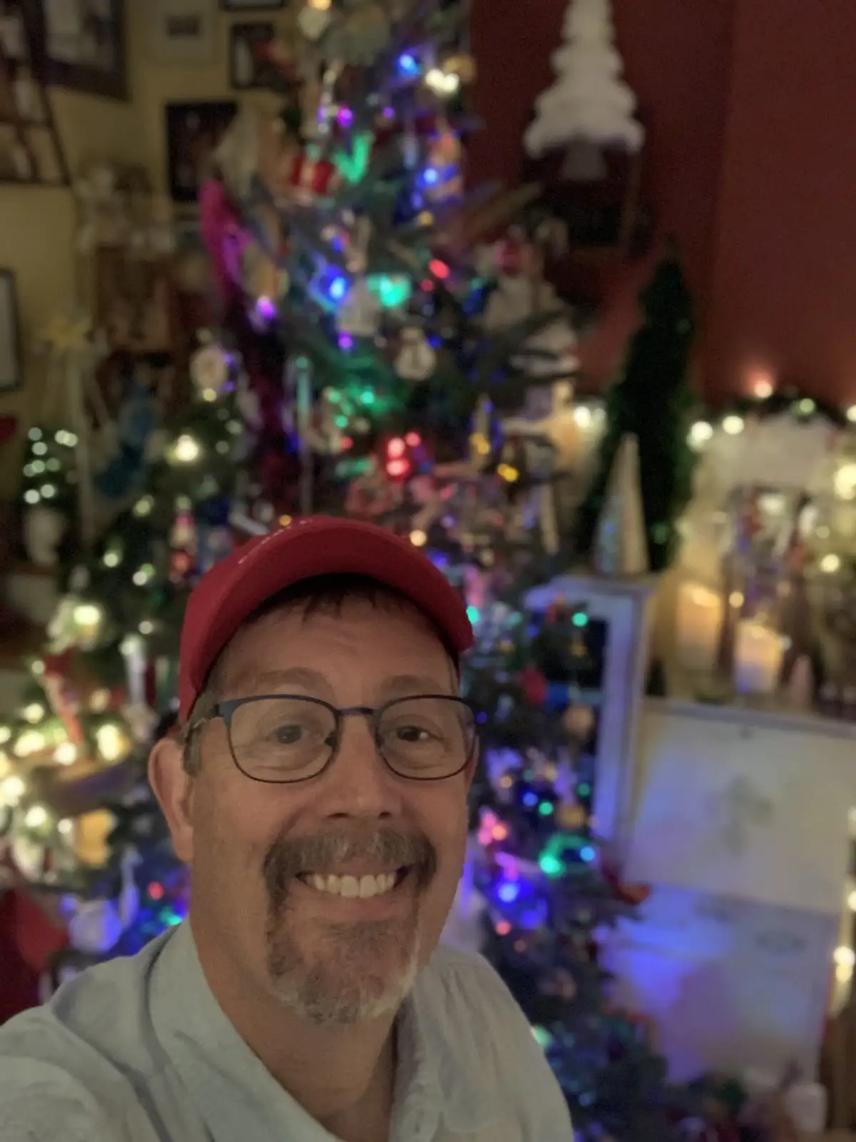 A man wearing a red cap and glasses smiles at the camera, with a brightly lit, decorated christmas tree and other holiday…