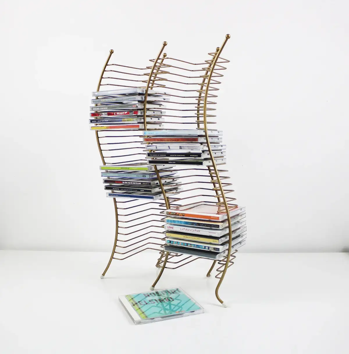A golden wire CD rack is visibly bent and warped under the weight of numerous CDs stacked on its shelves, with one CD case…