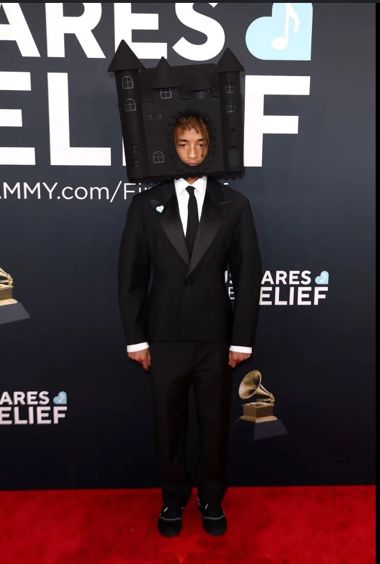 A man in a black tuxedo stands on a red carpet at a Grammy Awards event, wearing a large, handmade cardboard castle on his…