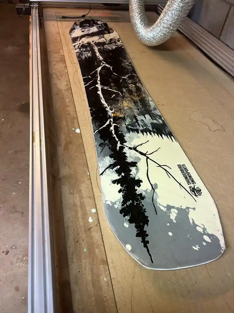 A snowboard with a black and white forest scene design lies on a workbench inside a machine, with a silver duct visible…