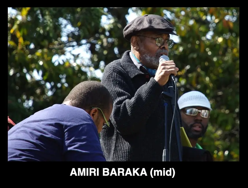 A man with a beard, glasses, and a cap speaks into a microphone outdoors, flanked by two other men. One man in a blue shirt…