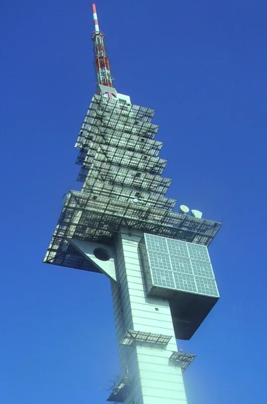 A low-angle photo of a tall, modern telecommunications tower against a clear blue sky, featuring a distinctive stepped…