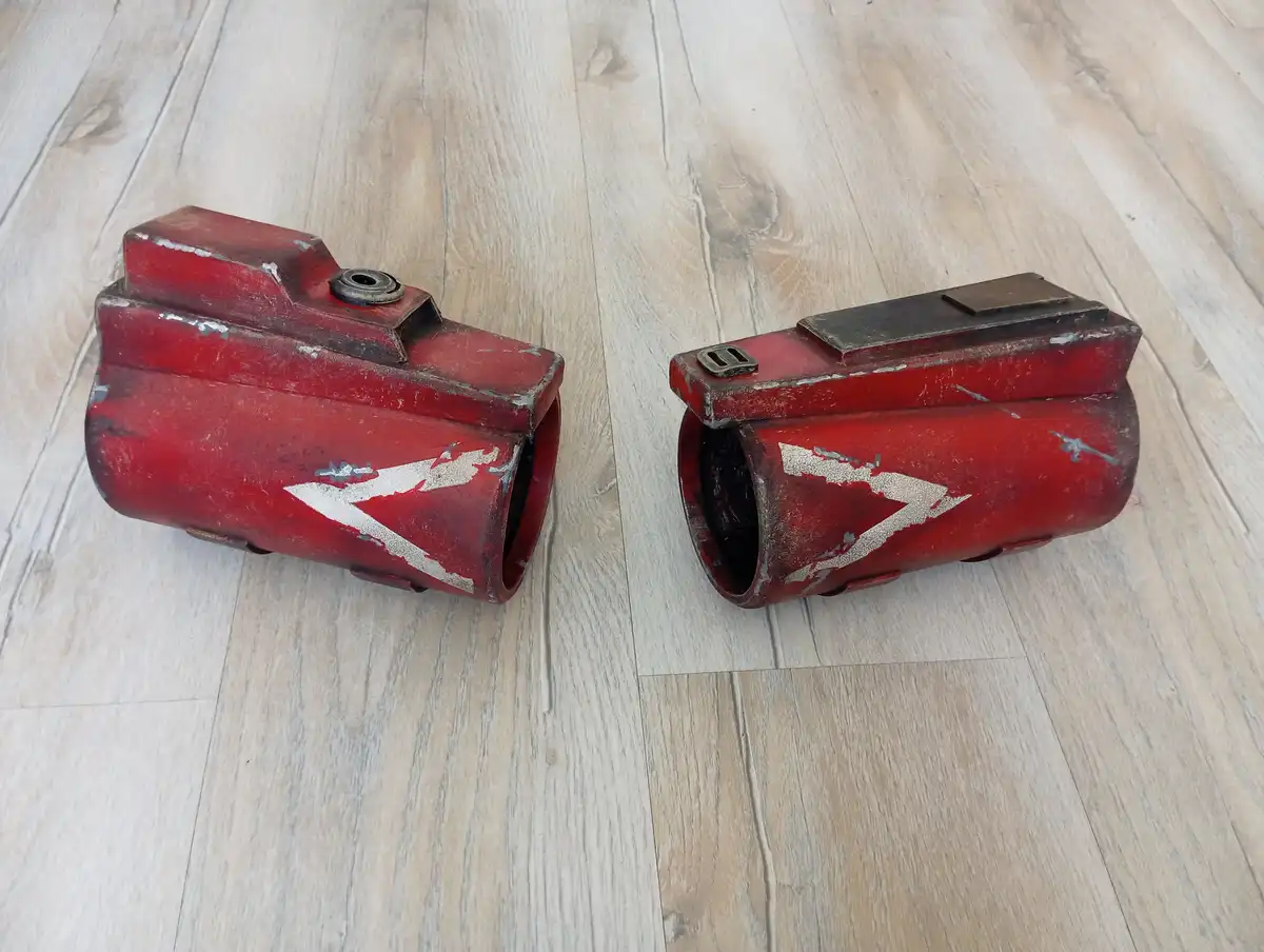Two red, weathered, cylindrical objects with white arrow decals lie on a light wood floor, one with a circular opening and…