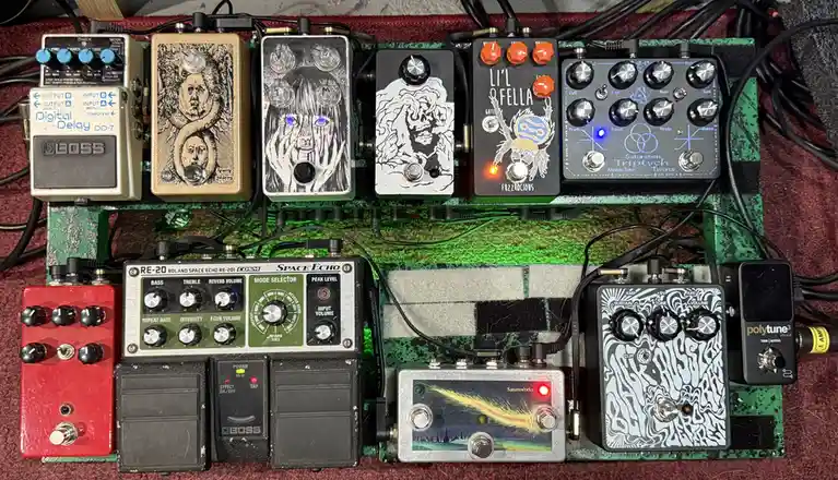 An overhead shot of a densely packed guitar pedalboard featuring numerous effects pedals with varied artwork, knobs, and…
