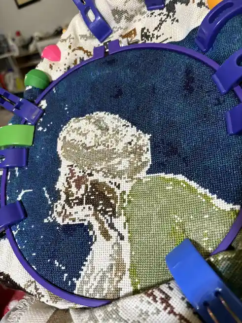 A close-up photo shows a partially completed cross-stitch project in a purple hoop, depicting a stylized animal figure…