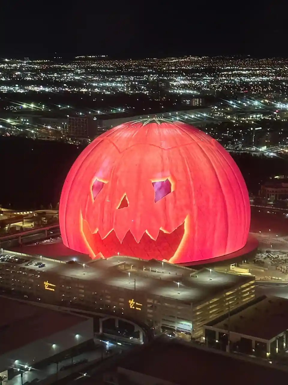 a nighttime aerial view shows a large, dome-shaped building illuminated to resemble a giant jack-o'-lantern with glowing…