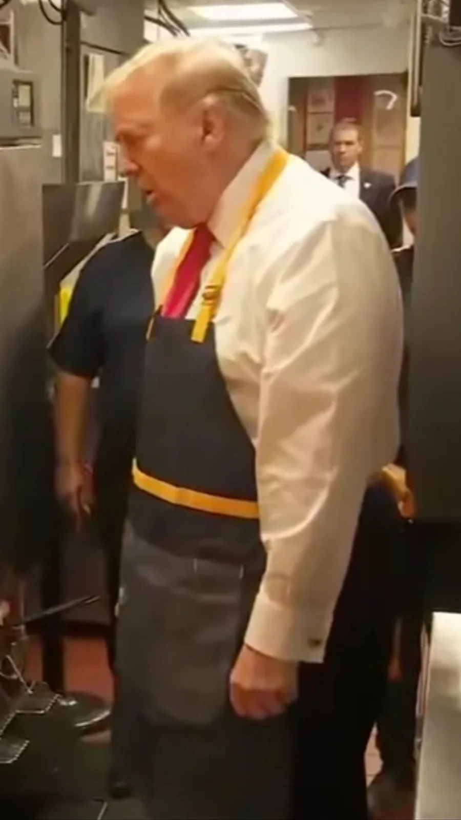 A man with blond hair wearing a white shirt, red tie, and a dark apron with yellow straps stands in what appears to be a…