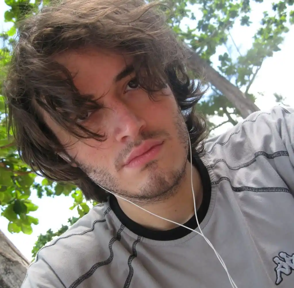 A young man with wavy brown hair and a light beard looks directly at the camera with a slightly bored or uninterested…