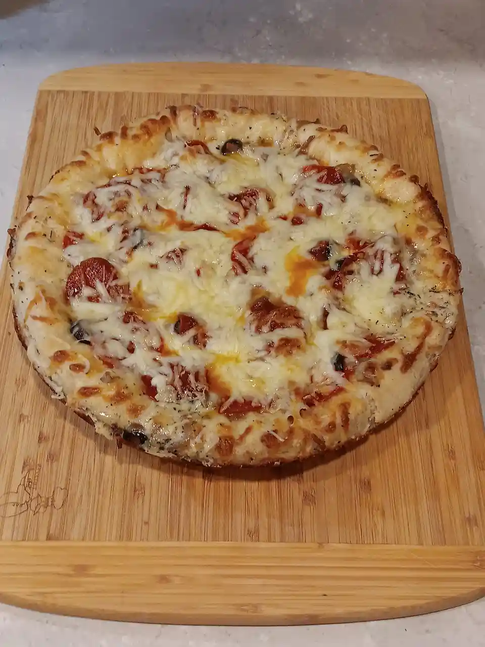 A homemade pepperoni and olive pizza with melted cheese sits on a wooden cutting board, looking perfectly cooked and…