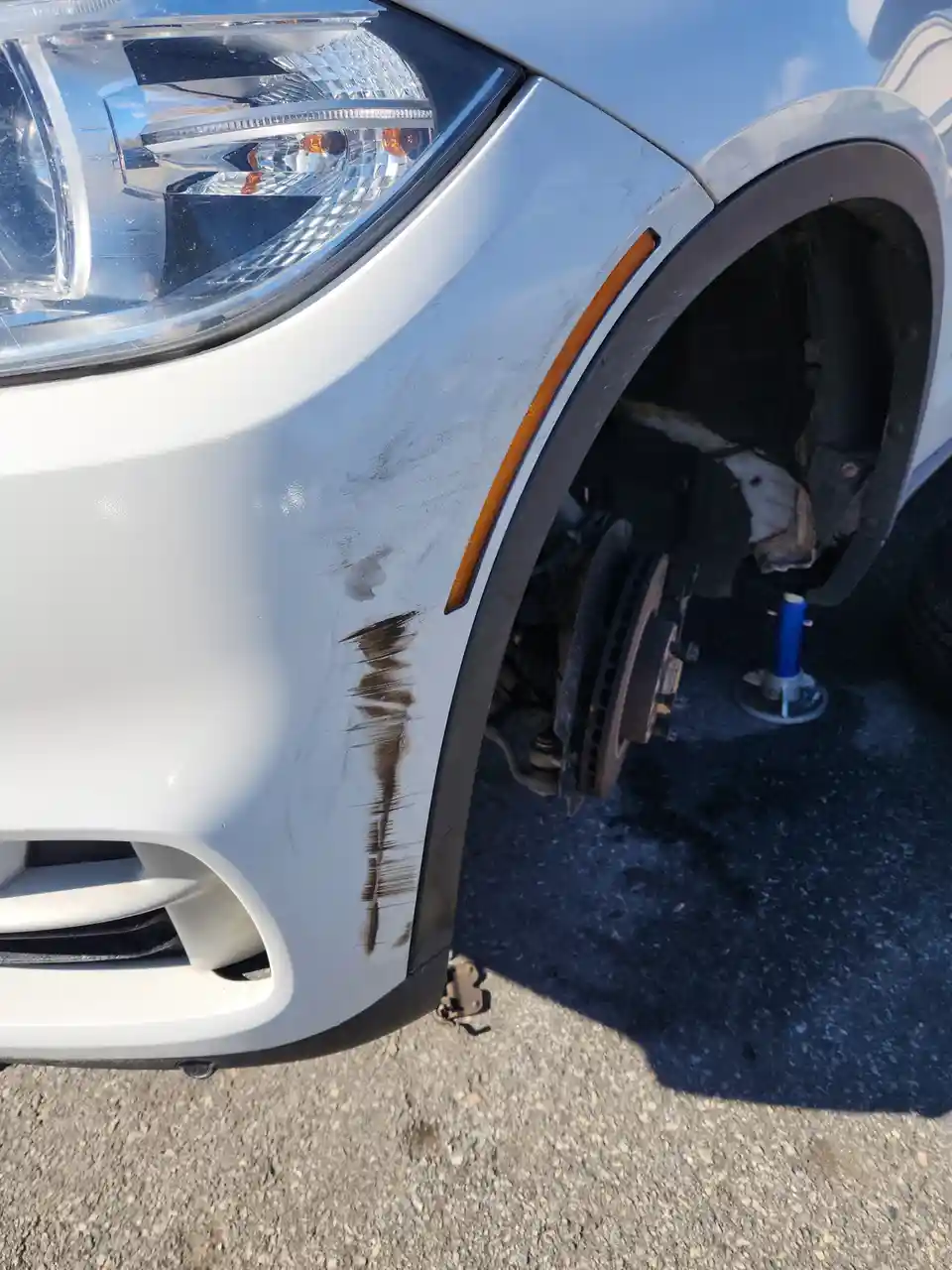 A close-up of a white car's front fender shows a large scrape and brown dirt streaks, with the wheel removed and the car…