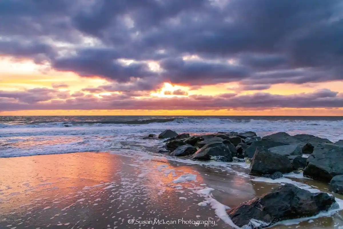 A serene beach scene at sunset with dramatic purple and orange clouds, waves crashing on rocks, and wet sand reflecting the…