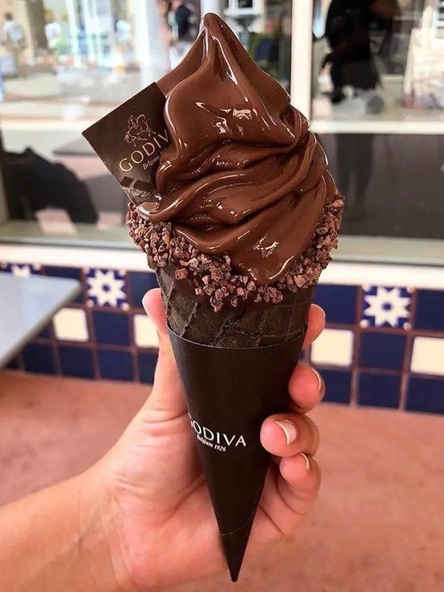 A hand holds a Godiva chocolate soft-serve ice cream cone, topped with chocolate sauce and chocolate shavings, with a Godiva…