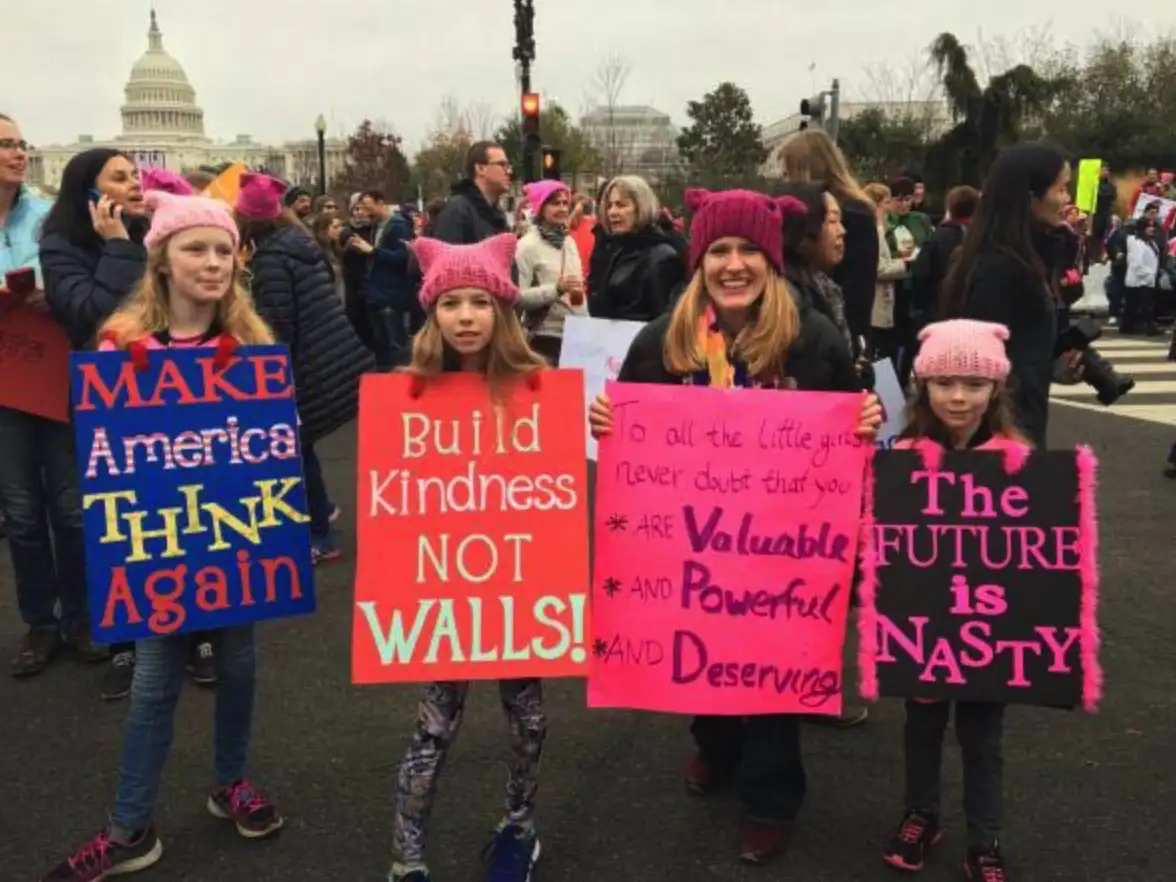Four young girls in pink cat-ear hats stand in a protest in front of the U.S. Capitol, each holding a handmade sign with…