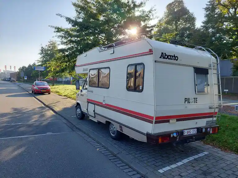 A white Abarto R 450 camper van with red stripes is parked on a paved curb next to a road, with the sun shining through…