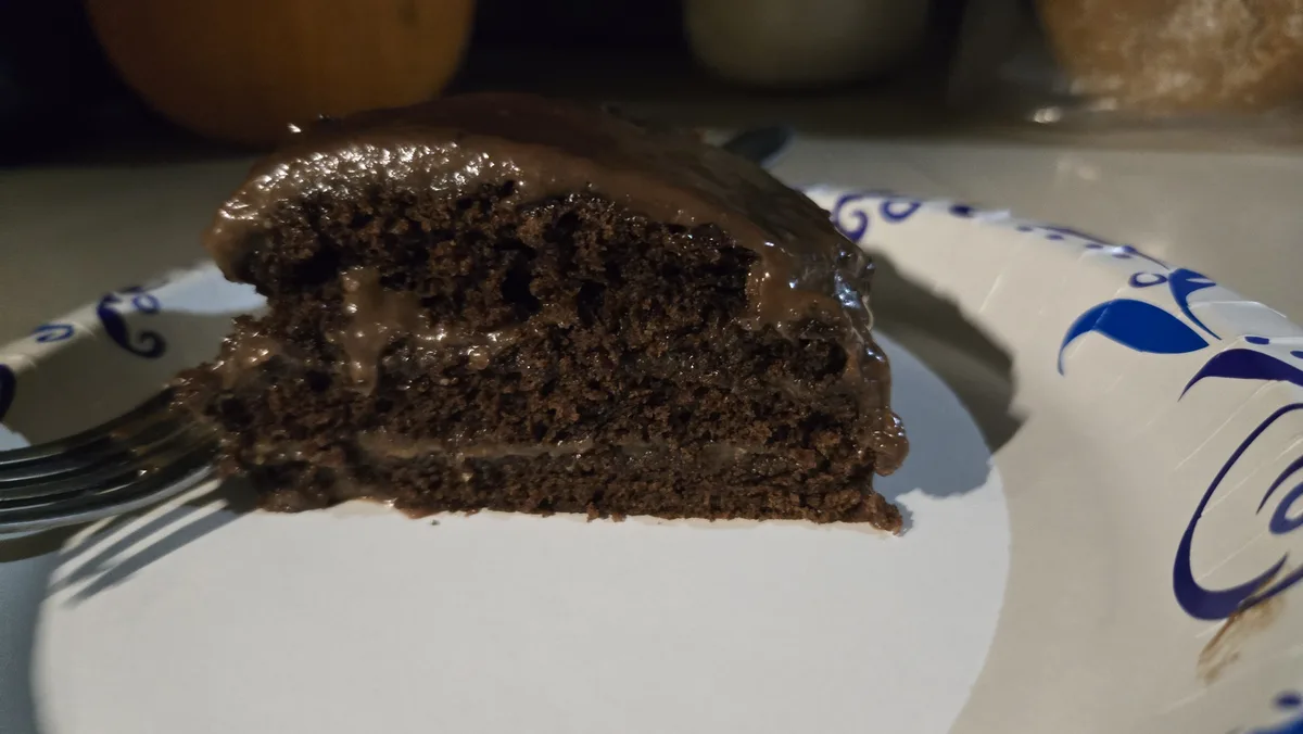 A close-up photo shows a slice of rich, moist chocolate cake with glossy chocolate frosting on a white paper plate with blue…
