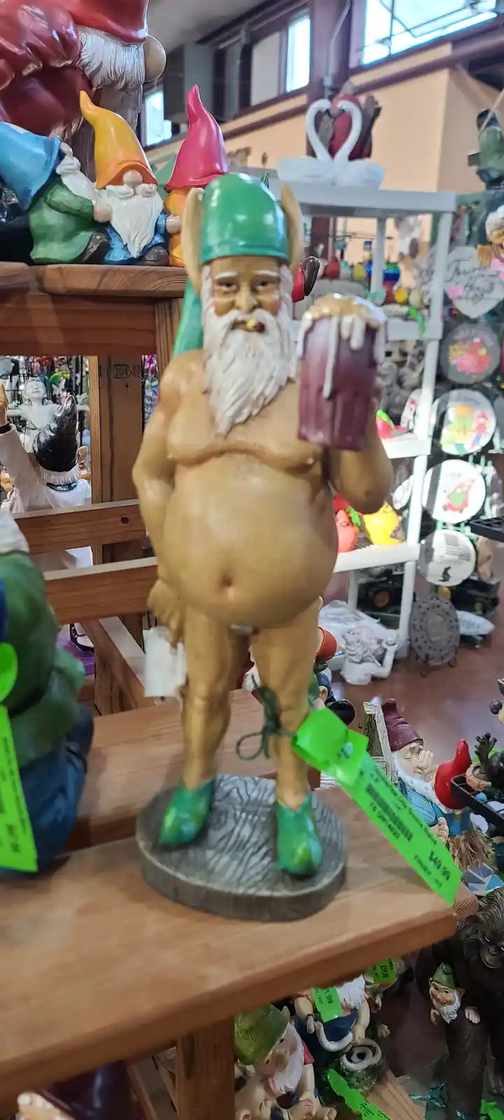 A large, shirtless garden gnome statue with a green horned helmet, a white beard, and green shoes stands on a wooden shelf…