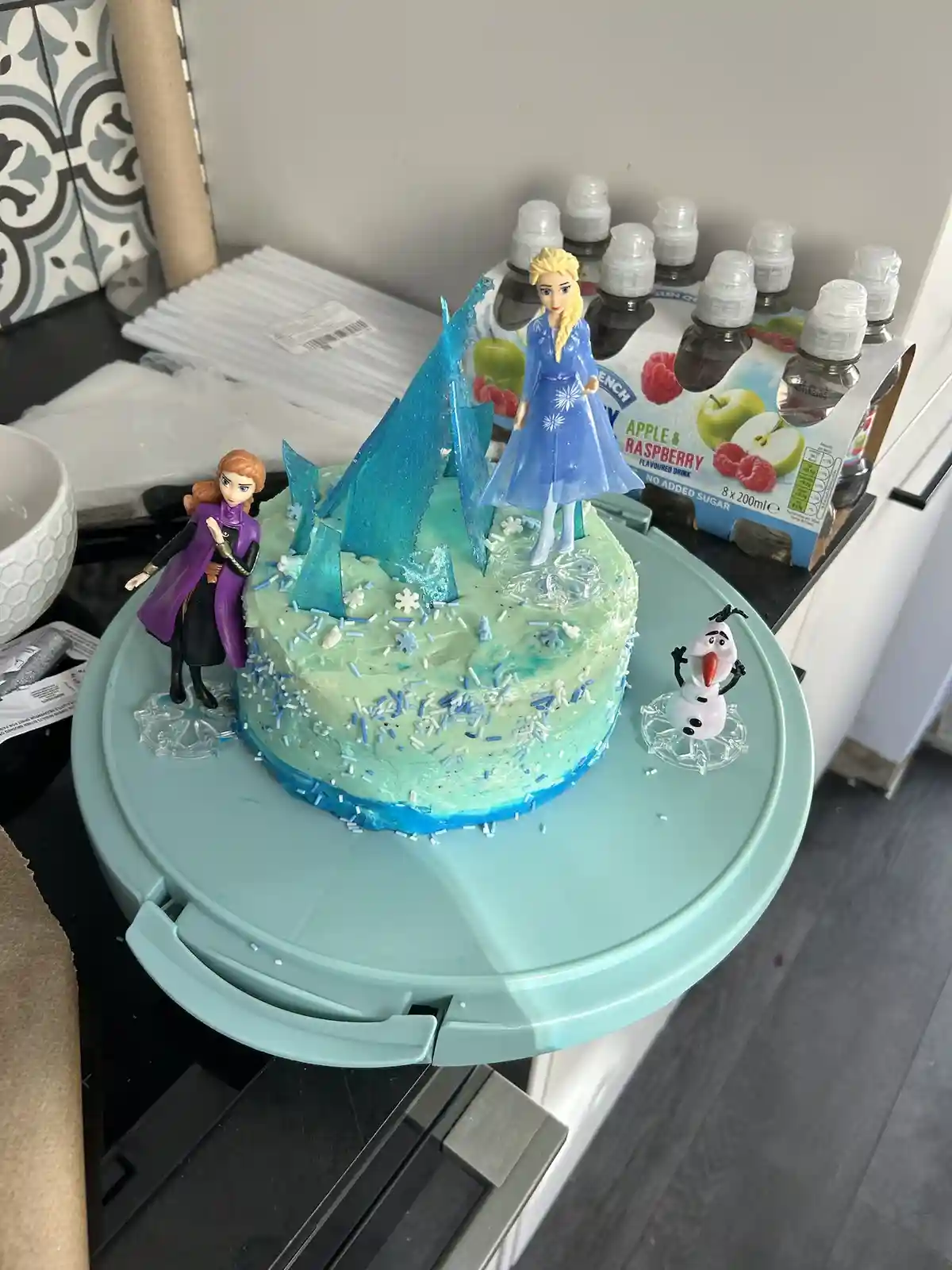 A light blue frosted birthday cake decorated with blue and white sprinkles and ice-like decorations sits on a light blue…
