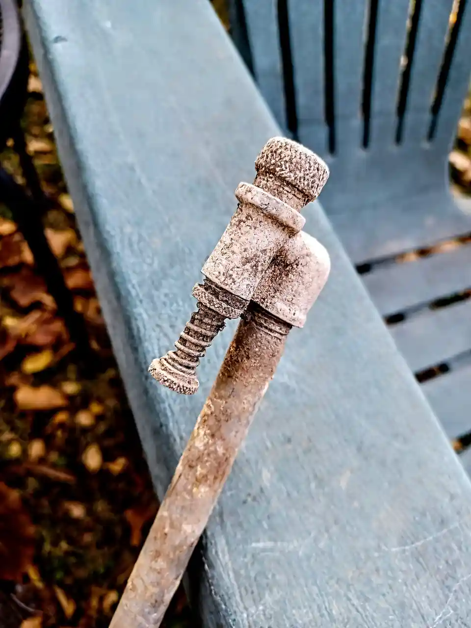 A rusty bolt and nut, covered in dirt and grime, are positioned upright on a weathered blue park bench, resembling a person…