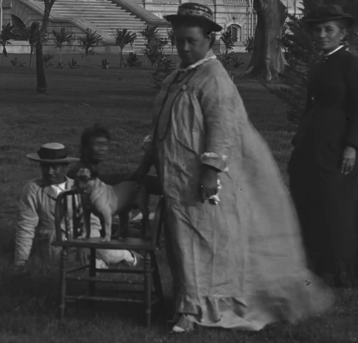 A vintage black-and-white photograph shows a woman in a long dress and hat standing in a grassy area with a building and…