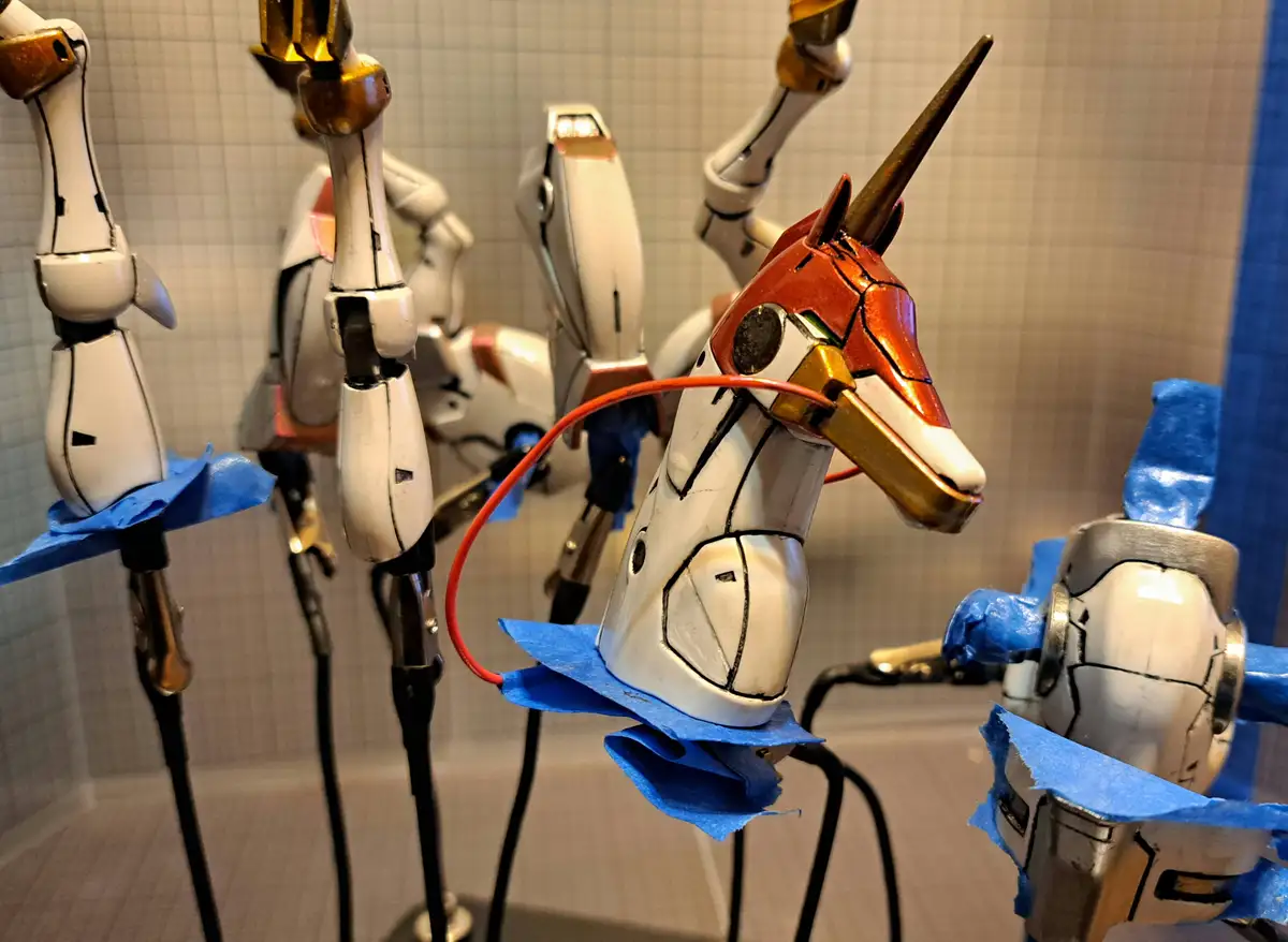A close-up photo shows several white and gold robot model heads, resembling mecha or anime characters, mounted on stands…
