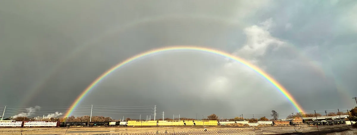 A wide-angle photo shows a vivid double rainbow arcing over a train yard with freight cars, power lines, and a chain-link…